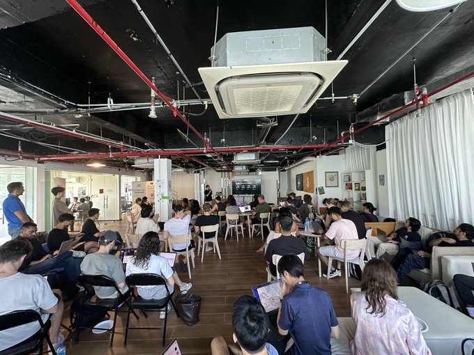The image shows a group of people sitting around tables in an office setting. There are at least 12 people visible in the scene, some of them wearing backpacks or carrying handbags. The chairs they're sitting on appear to be black and white. A large screen is mounted on the ceiling above the group, which could be used for presentations or meetings.

The post that seems to be related to this image reads: "If you build with Virtuals, @base will support you". This suggests that the office space might be designed to accommodate virtual meetings and events using a virtual meeting platform like Google Meet or Zoom. The presence of multiple people sitting around tables indicates that they are engaged in discussions or collaborative work activities.