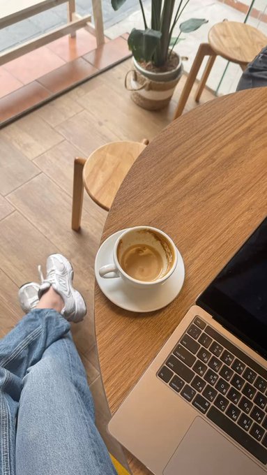 A first-person view of someone sitting at a wooden café table with an espresso cup, a MacBook laptop, and their feet in white sneakers and jeans visible below.
