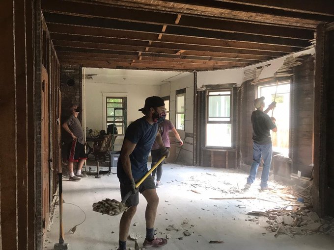Workers demolish the interior wall and ceiling of an old building, with debris scattered across the floor and exposed studs and beams visible.