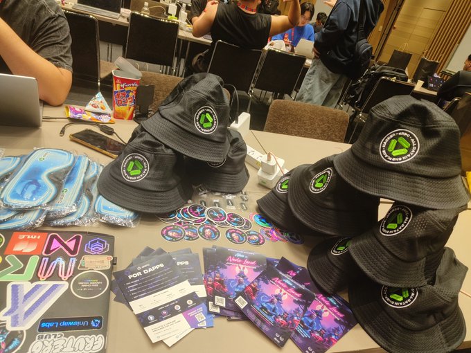 A table display features stacks of black bucket hats with a green recycling logo reading 'New York Linux Users Group,' alongside stickers, stickers, patches, and event flyers for 'Pancake Breakfast' and 'Pork Papers.'