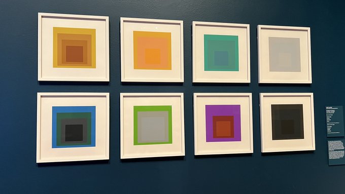 A gallery wall displaying eight framed abstract prints of nested colored squares in various hues (orange, teal, gray, blue, green, purple, black), with informational placards visible on the right.