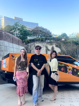 Three people posing in front of an orange Lamborghini SUV outdoors on a sunny day, with the central man wearing a black MM shirt.
