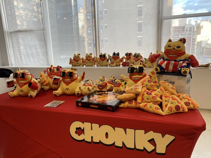 A promotional table covered with a red 'CHONKY' branded tablecloth displays various Chonky cat plush toys wearing sunglasses and pizza-shaped cushions, with a city view in the background.