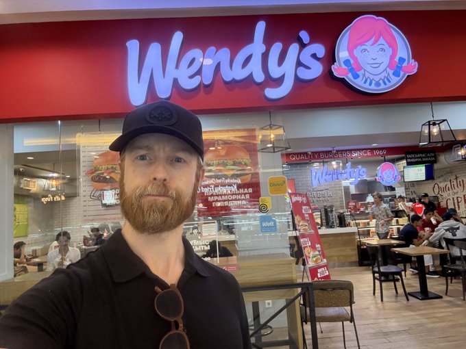 A man wearing a black cap and shirt poses for a selfie in front of a Wendy's restaurant, with the iconic red "Wendy's" logo and the slogan "Quality is our recipe" visible in the background, capturing a casual visit that aligns with the "boots on the ground $WEN" post.