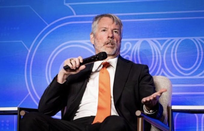 A man with gray hair and a beard, wearing a black suit with an orange tie, sits on a stage holding a microphone while gesturing with his other hand during what appears to be a speaking engagement.

The image relates to the post as a visual accompaniment to commentary on Bitcoin's price movement, with the speaker likely discussing the "BTC64,000 Dollar Battle" and the three key factors mentioned in the post that are driving the current market.