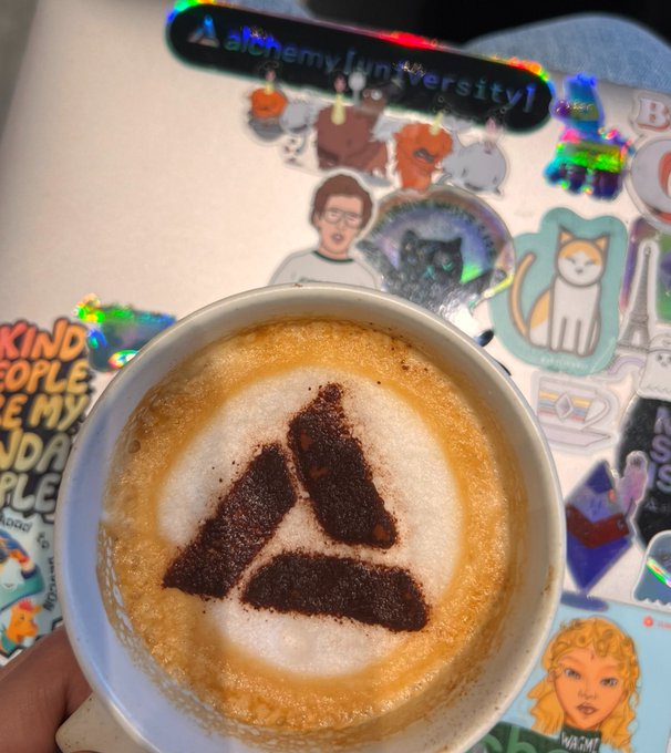A cappuccino featuring an "A" logo in cinnamon/chocolate powder sits on a laptop covered in stickers, including one reading "alchemy [university]," visually representing the celebration of Alchemy's engineering team achievements highlighted in the post.