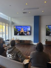 People sit on couches watching a television mounted on a blue and white wall in what appears to be a lounge or waiting area.