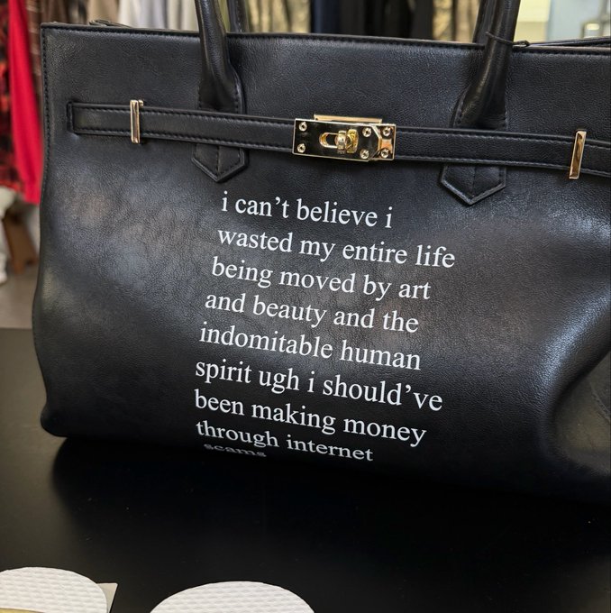 A black leather Birkin-style handbag with gold hardware displays white printed text: "i can't believe i wasted my entire life being moved by art and beauty and the indomitable human spirit ugh i should've been making money through internet."