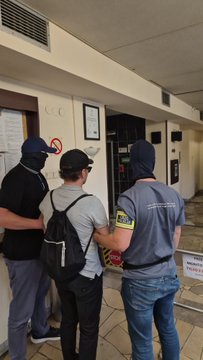 Photograph showing two uniformed officers escorting a handcuffed man in a white shirt and backpack through a building corridor.