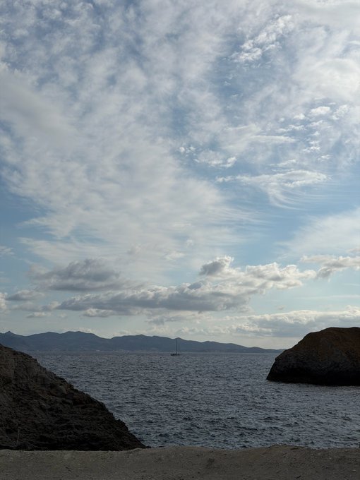 A scenic view of a calm body of water bordered by rocky cliffs, with mountains and a lone sailboat in the distance under a sky filled with scattered clouds.