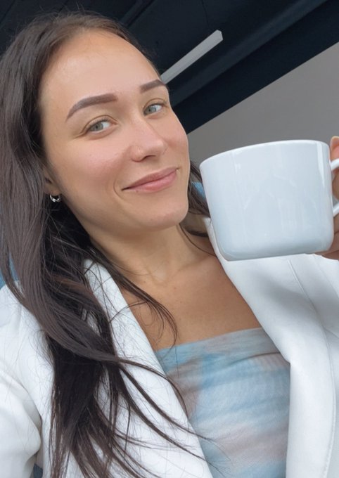 A smiling woman with long dark hair, wearing a white jacket over a blue top, holds up a large white ceramic mug.