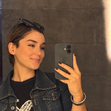 A young woman with short hair and sunglasses on her head takes a mirror selfie with her iPhone, smiling softly while wearing a denim jacket.
