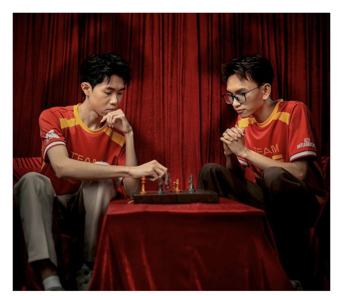 Two young men in red and yellow 'TEAM' jerseys intensely focused on a game of chess at a red velvet-covered table against red curtain backdrop.