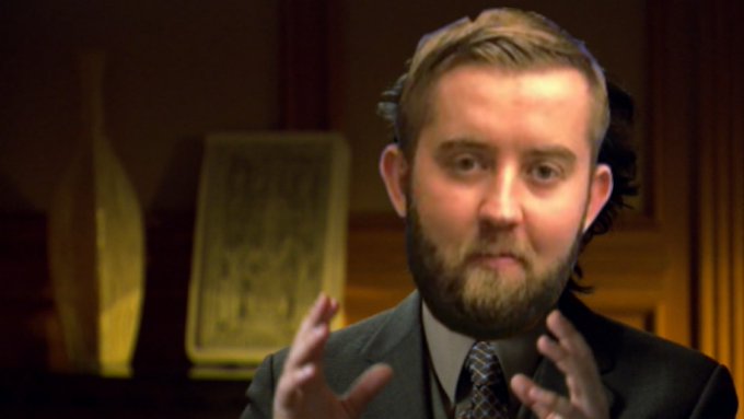 A bearded man in a dark suit and tie gestures with both hands raised while speaking, set against a blurred warm-toned interior background with a decorative object.