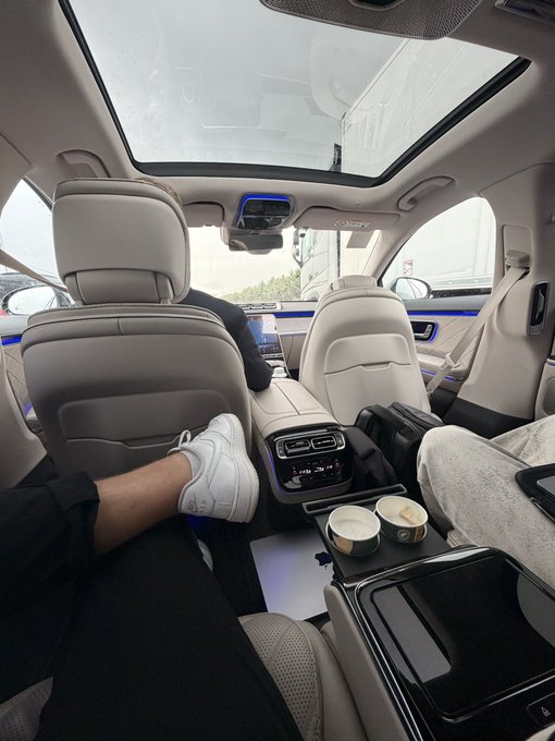 The image shows a luxurious car interior with a panoramic sunroof, where a passenger is relaxing in the back seat with their feet up on a center console holding two coffee cups, while the driver appears focused on the road ahead. This aligns with the post celebrating crypto market gains and a sponsored ride to the Dutch Grand Prix at Zandvoort, with Crypto.com likely providing the premium transportation.