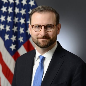 A portrait of a man in a suit and blue tie with glasses, standing in front of an American flag.
