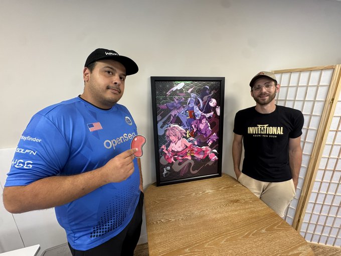 Two men pose next to a framed anime poster on a wooden table; one wears a blue esports jersey with 'OpenServ' and a US flag, while the other wears a black 'INVITATIONAL AKM TCM 2024' t-shirt.