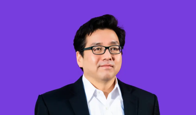 A portrait of an Asian man wearing glasses and a dark suit jacket over a white shirt, photographed against a solid purple background.