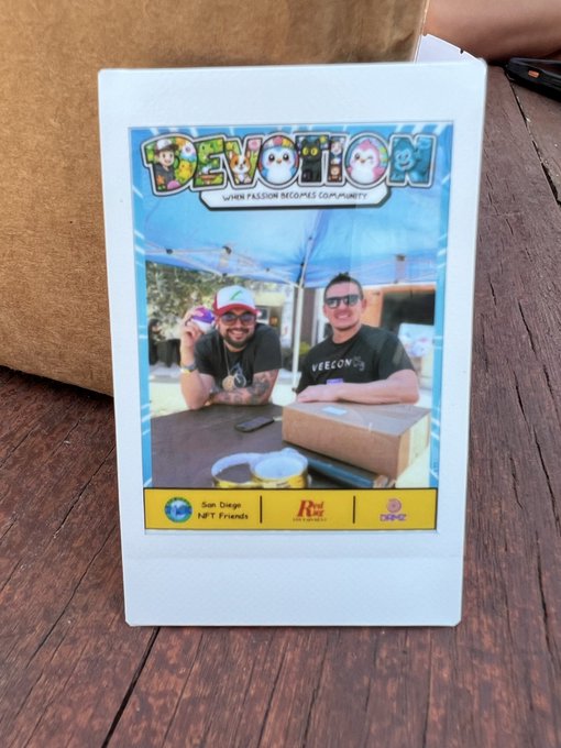 A custom Polaroid photo sits on a wooden table, featuring two smiling men under a tent with a colorful "DEVOTION" banner reading "When Passion Becomes Community," and includes a yellow footer with sponsor logos (San Diego NFT Friends, Art, and Devotion). The photo captures a fun, community-oriented moment, perfect for sharing on social media as the post encourages recipients to do.