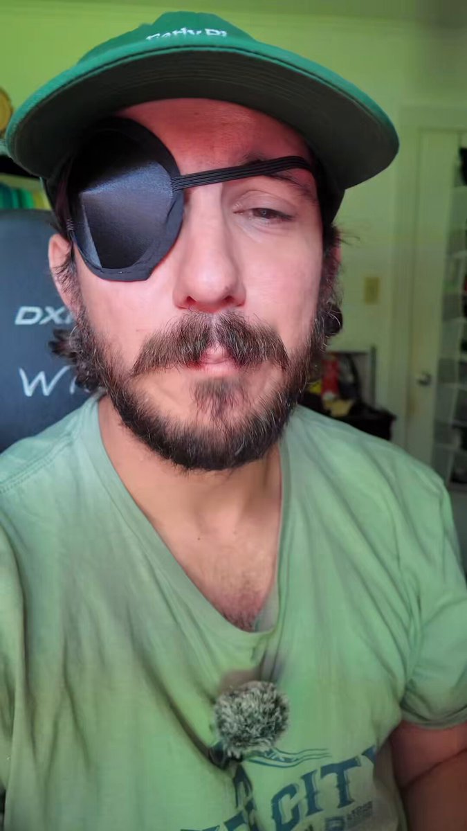 The image features a man wearing a green t-shirt, a black baseball cap with a white eye patch, and a green shirt underneath his hat. The man has a beard and mustache, giving him a distinctive appearance. He appears to be posing for a photo or participating in an online gaming event.