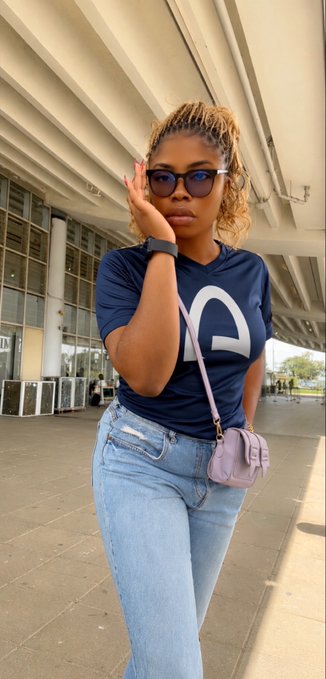 A stylish woman wearing glasses, a navy blue t-shirt, light blue jeans, and carrying a lavender crossbody bag, posing outdoors under a modern architectural overhang.
