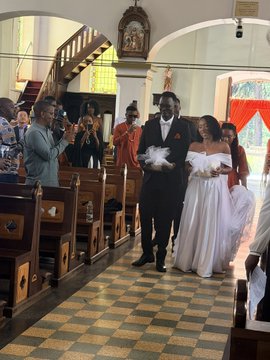 A wedding scene inside a church where a bride in a white dress and groom in a dark suit walk down the aisle, accompanied by attendants and followed by a photographer documenting the moment.
