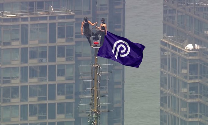 Two people are perched atop a cellular tower waving a purple Polkadot (DOT) flag, with high-rise buildings visible in the background.