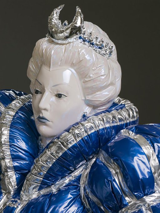 A sculptural figure with a white ceramic face wearing a tiara and a silver crescent moon ornament, adorned in an elaborate puffy outfit made of shiny blue and silver metallic fabric.