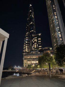 A nighttime view of an illuminated skyscraper and modern city architecture with people walking along a waterfront promenade.