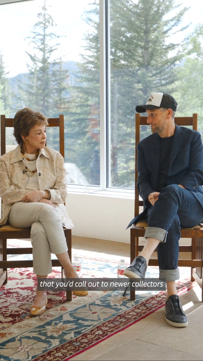 An older woman in a cream embellished jacket sits across from a man in a navy blazer and baseball cap, having a discussion in a sunlit room with large windows showing pine trees. The caption reads "that you'd call out to newer collectors," referencing her advice about floor prices for new art collectors.