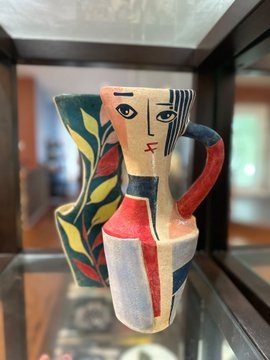 A cubist-style ceramic pitcher features a stylized face with a long nose and bold green, red, yellow, and black geometric patterns.