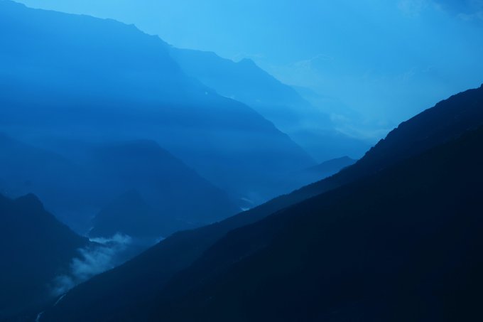 Atmospheric blue mountain landscape with layered peaks shrouded in mist and low-hanging clouds.