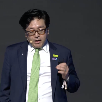 A bespectacled man in a navy suit and green tie speaks on stage at Korea Blockchain Week 2026, delivering a presentation that aligns with the caption about Tom Lee, Chairman of Bitmine (ticker: BMNR), predicting the largest bull market cycle yet.