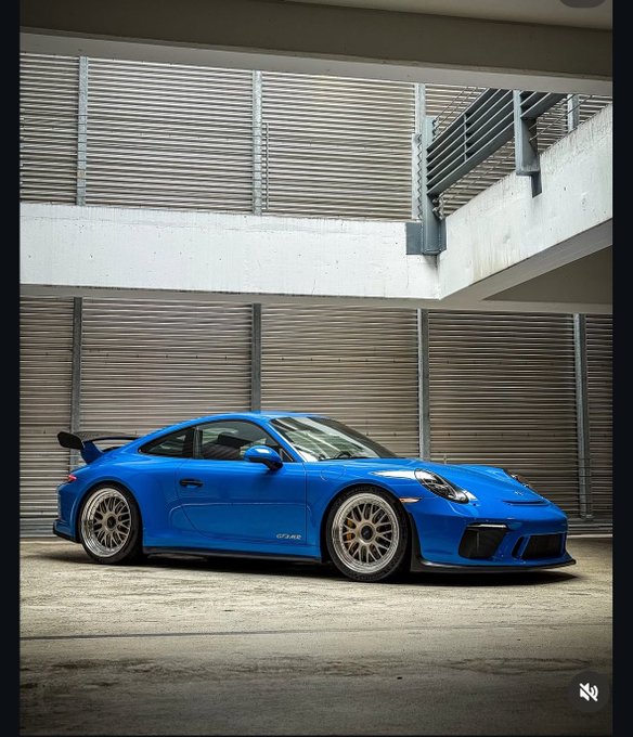 A blue Porsche 911 (991.2) GT3 MR in PTS Voodoo Blue is parked in front of a modern industrial building, showcasing its BBS wheels and "GT3 RS" side decal as advertised in the off-market sale listing.