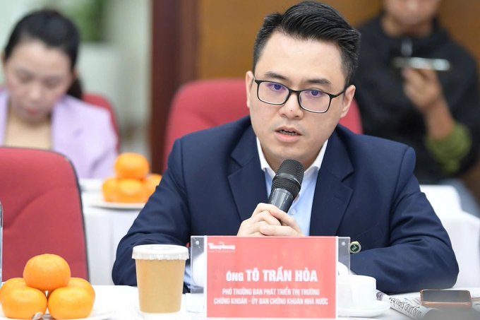 A man in a suit speaks into a microphone at a panel discussion, identified by the name placard as "Ông Tô Trần Hòa, Phó Trưởng Ban Phát Triển Thị Trường Chứng Khoán - Ủy Ban Chứng Khoán Nhà Nước" (Deputy Head of Market Development Department - State Securities Commission of Vietnam), with oranges and a coffee cup on the table before him. The image illustrates the accompanying post about his announcement regarding the upcoming Vietnam asset exchange, which will only support VNĐ trading pairs and list 5 types of assets.