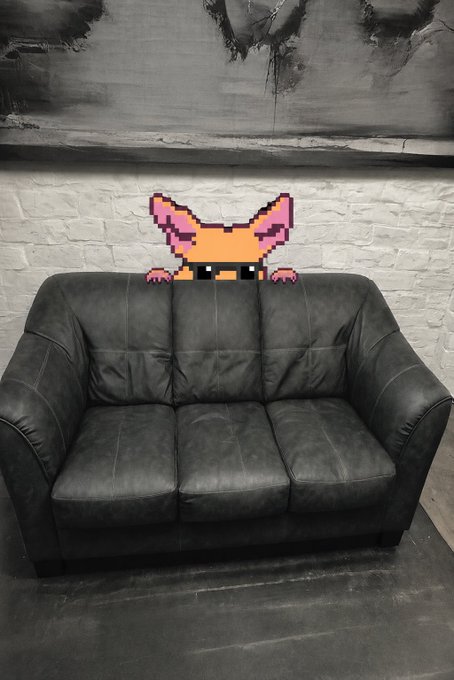 A pixel art piece of a cat peeking over the back of a black leather couch against a distressed wall.
