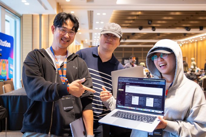 Three smiling people pose together at what appears to be a tech conference, with the person on the right holding an open laptop displaying a software interface.
