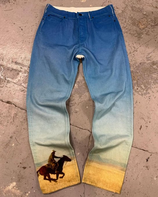 A pair of custom-designed jeans displayed on a stone surface, featuring a gradient ombre dye effect that transitions from deep blue at the waist to pale yellow at the ankles, with a hand-painted illustration of a cowboy on horseback near the left hem.