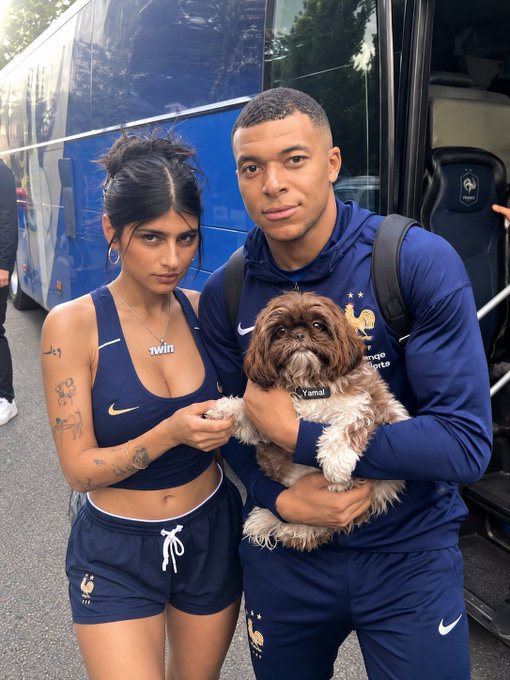 Kylian Mbappé, wearing a French national team tracksuit, stands next to a woman in matching France athletic wear while holding a small dog named 'Tymal' in front of a team bus.