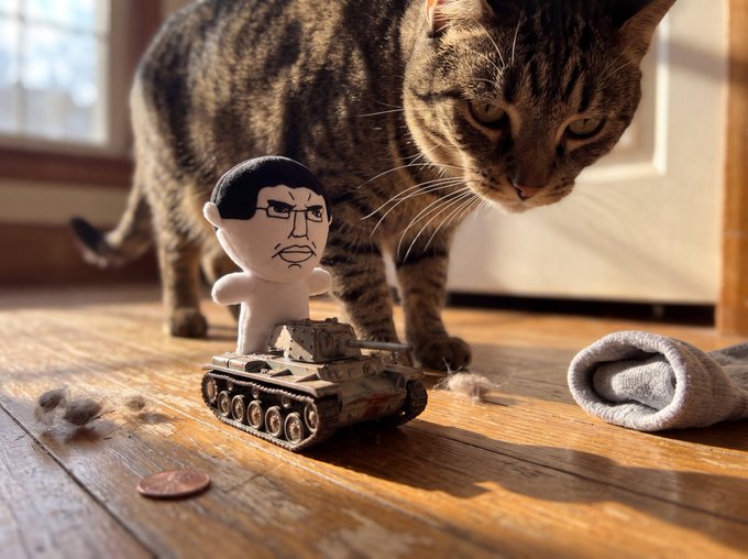 A tabby cat curiously inspects a small toy tank topped with a bobblehead figure on a wooden floor next to a penny and a rolled-up sock.
