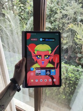 A hand holding a tablet displaying an animated elf character with blonde hair and green outfit, set against a background of lush green foliage viewed through a window.
