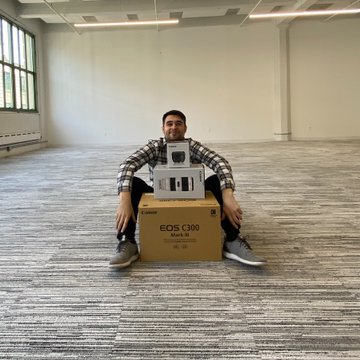 A man sits on the floor of a large empty office space, posing behind a cardboard box labeled "EOS C300 Mark III" with the camera displayed on top, symbolizing the significant investment in production equipment that marked the launch of his media venture four years ago.