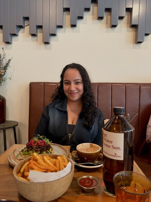 A young woman with curly hair wearing a denim jacket, sitting at a restaurant table with a salad, sweet potato fries, and a bottle of 'Water from the Sun' tea.