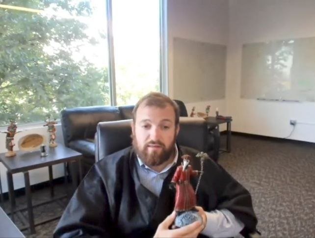 A man with a beard wearing a black robe sits in an office, holding a small figurine of an angel or saint.