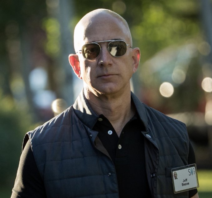 A bald man wearing sunglasses and a dark vest over a polo shirt, with an ID badge clipped to his vest reading 'Jeff Bezos'.