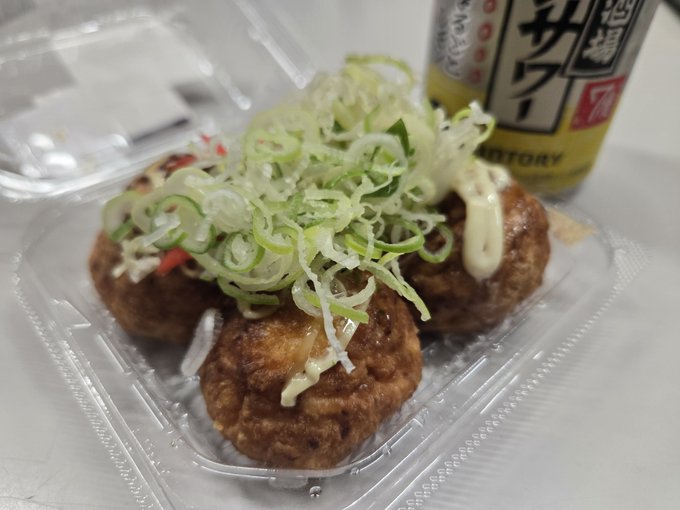 A plastic takeout container holds takoyaki (Japanese octopus balls) topped with shredded green onions and mayonnaise, accompanied by a can of Suntory Kinmugi barley tea.