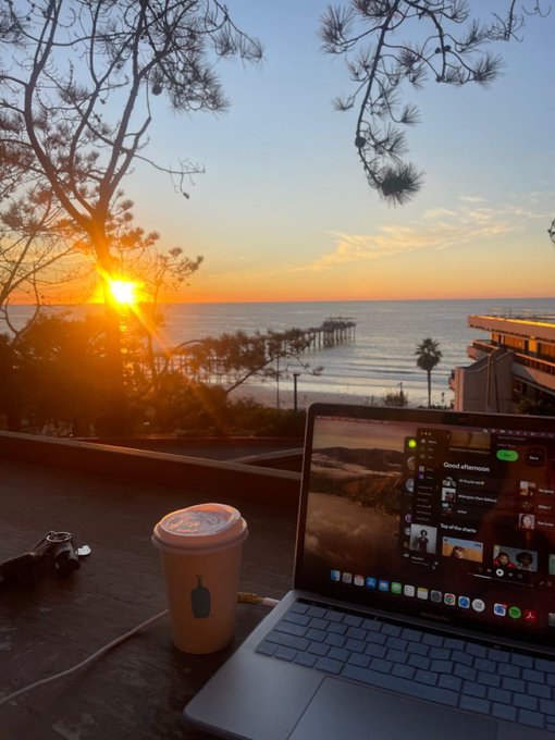 A coffee and laptop sit on an outdoor table overlooking a stunning coastal sunset with a pier in the distance, capturing the relaxed "Good afternoon" vibe of a Friday work session. The Spotify playlist visible on the screen and the scenic ocean view perfectly match the post's casual GM energy and remote-work lifestyle.