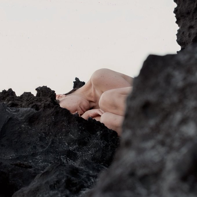 The image shows a person lying down on rocks, with their head resting against a rock face. They are not wearing any clothing or accessories, which adds an element of rawness to the scene. The photo appears to be taken from behind the person, giving a sense of depth and perspective. The caption "Good morning Self portrait 0.07 ETH" suggests that this image was captured during the early hours of the day when the sun is rising, adding a serene atmosphere to the scene.