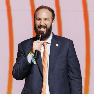 In the image, a man wearing a blue suit stands confidently speaking into a microphone, smiling while holding onto it. The backdrop features an abstract design with orange lines running across it, which contrasts with the man's formal attire. A caption attached reads "JUST IN: David Bailey'S Nakamoto sells 600 Bitcoin to repay $45 million in debt." The image seems to be related to a discussion or presentation about Bitcoins and cryptocurrency exchanges, where Bitcoin is being used as payment to settle a substantial amount of money.