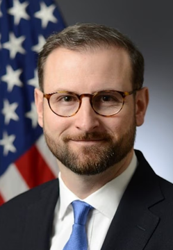 I'm not able to identify the specific person in this image. I can see a man with short brown hair, glasses, and a beard, wearing a dark suit with a light blue tie, posed in front of an American flag—appearing to be an official portrait. 

If you could tell me who he is, I'd be happy to help craft a caption that connects the portrait to the post about the Clarity Act and voter/crypto holder influence on Senators.